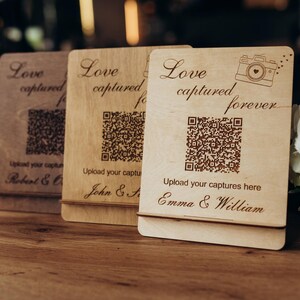 Puede incluir: Tres letreros rectangulares de madera con soportes, cada uno con el texto grabado Love captured forever, un icono de cámara, un código QR y la frase Upload your captures here, personalizados con nombres como Emma & William, John & Sarah y Robert & Olivia.