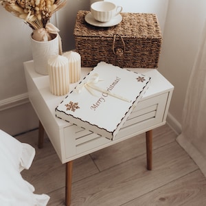 Op de afbeelding: Een rechthoekige witte houten doos met een geschulpte rand, voorzien van de tekst Merry Christmas in bruine letters, twee bruine sneeuwvlokpictogrammen en een cr&egrave;mekleurig lint gestrikt aan de bovenkant, op een wit nachtkastje.