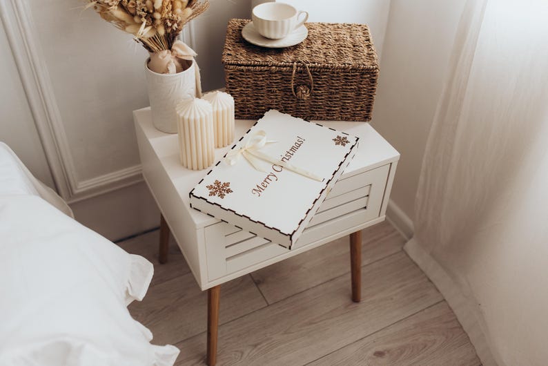 Op de afbeelding: Een rechthoekige witte houten doos met een geschulpte rand, voorzien van de tekst Merry Christmas in bruine letters, twee bruine sneeuwvlokpictogrammen en een cr&egrave;mekleurig lint gestrikt aan de bovenkant, op een wit nachtkastje.