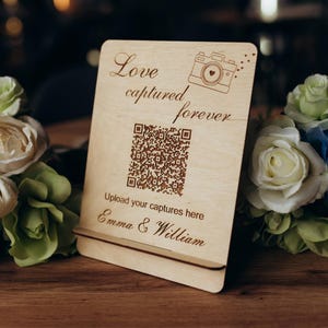 May include: A wooden sign engraved with "Love captured forever" and a camera icon. It features a QR code and the text "Upload your captures here Emma & William". The sign is set amongst white and blue floral arrangements.