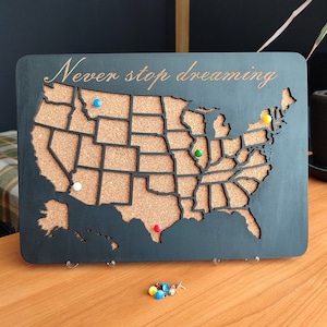 Pode incluir: Um quadro retangular de madeira preta com um mapa de cortiça dos Estados Unidos com as fronteiras estaduais. O texto "Never stop dreaming" está gravado no topo. Vários percevejos coloridos estão inseridos no mapa, com mais abaixo.