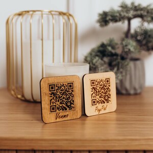 May include: Two square light-colored wooden signs with rounded corners, each featuring a laser-engraved black QR code. The left sign displays the text Venmo, and the right sign displays the text PayPal in a cursive font below the QR code.