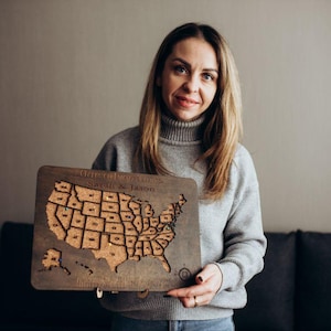 Op de afbeelding: Een rechthoekige houten kaart van de Verenigde Staten met een kurken inleg die de contouren van de staten toont. Het bord is aan de bovenkant gegraveerd met de tekst Our Adventures en Sarah & Jason, en onderaan met extra tekst, met kleine spelden.