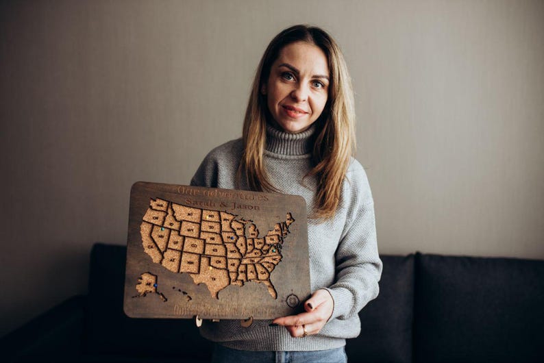 Op de afbeelding: Een rechthoekige houten kaart van de Verenigde Staten met een kurken inleg die de contouren van de staten toont. Het bord is aan de bovenkant gegraveerd met de tekst Our Adventures en Sarah & Jason, en onderaan met extra tekst, met kleine spelden.