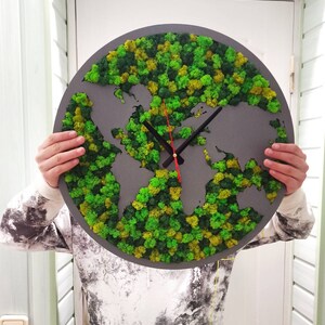 Op de afbeelding: Een ronde grijze wandklok met een wereldkaartontwerp. De kaart is gevuld met groen en geel mos, en de wijzers zijn zwart met een rode secondewijzer. De klok wordt door een persoon vastgehouden.