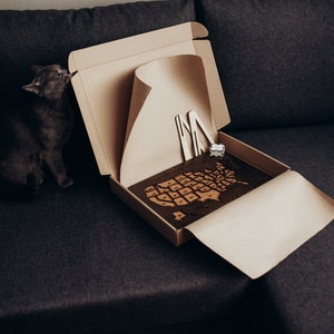 Op de afbeelding: Een kartonnen verzenddoos met een donkerbruine houten puzzel van de kaart van de Verenigde Staten. De doos bevat twee lichtgekleurde houten steunen en een kleine witte kubus. Een grijze kat zit naast de open doos.