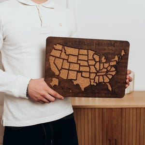 Op de afbeelding: Een rechthoekig houten wandbord met een kurkkaart van de Verenigde Staten van Amerika. De donkerbruine houten lijst omringt de kurken inleg, die met een laser is uitgesneden in de vormen van de vijftig staten. De tekst Never Stop Exploring is bovenaan gegraveerd.
