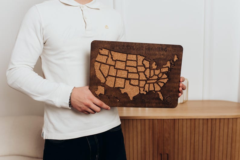 Op de afbeelding: Een rechthoekig houten wandbord met een kurkkaart van de Verenigde Staten van Amerika. De donkerbruine houten lijst omringt de kurken inleg, die met een laser is uitgesneden in de vormen van de vijftig staten. De tekst Never Stop Exploring is bovenaan gegraveerd.