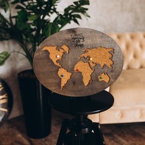 May include: A wooden world map with cork surface and a quote "Collect moments not things" on the top. The map is mounted on a black tripod stand. There are colorful push pins on the map.