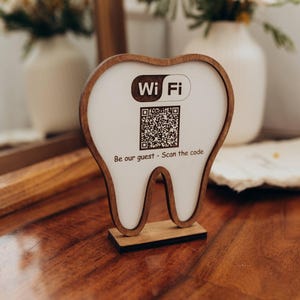 Op de afbeelding: Een houten Wi-Fi-bord in de vorm van een tand. Het bord heeft een wit midden met een QR-code en de woorden "Wi Fi" en "Be our guest - Scan the code" in bruin. Het staat op een houten oppervlak.