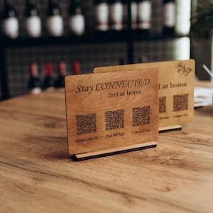 May include: Two rectangular light brown wooden signs with dark brown laser-engraved text and QR codes. The front sign features the text Stay CONNECTED feel at home above three QR codes labeled Guest Wi-Fi, Our MENU, and Contact Us. Each sign stands on a thin black base.