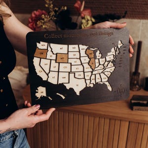 May include: A wooden map of the United States with state outlines. The map is dark brown with lighter brown states highlighted. The text "Collect moments, not things" is engraved at the top. The map is being held by a person.