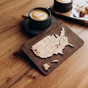 May include: A rectangular dark wood plaque featuring a light wood cutout of the United States map. The map includes engraved state borders, two small heart icons connected by a dashed path, and the text I wish I could teleport to you.