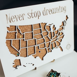 Op de afbeelding: Een wit rechthoekig houten bord met een uitgesneden kaart van de Verenigde Staten met een kurken achterkant. De tekst Never stop dreaming staat in bruin bovenaan gedrukt. Verschillende gekleurde punaises zitten in de kurkkaart.