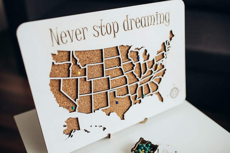Op de afbeelding: Een wit rechthoekig houten bord met een uitgesneden kaart van de Verenigde Staten met een kurken achterkant. De tekst Never stop dreaming staat in bruin bovenaan gedrukt. Verschillende gekleurde punaises zitten in de kurkkaart.