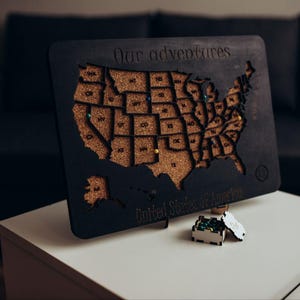 Op de afbeelding: Een zwarte houten kaart van de Verenigde Staten van Amerika met een kurken inleg. De tekst "Our adventures" is bovenaan gegraveerd en "United States of America" onderaan. Verschillende gekleurde punaises zitten in de kurken staten.