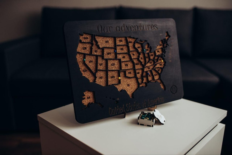 Op de afbeelding: Een zwarte houten kaart van de Verenigde Staten van Amerika met een kurken inleg. De tekst "Our adventures" is bovenaan gegraveerd en "United States of America" onderaan. Verschillende gekleurde punaises zitten in de kurken staten.