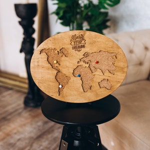 May include: A circular wooden world map with a cork centre and pins marking locations. The phrase "Never Stop Dreaming" is above the map. The map is displayed on a black stand.