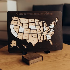 May include: Wooden map of the United States with state outlines and text "Collect moments, not things." The map is dark brown with lighter wood states. A small wooden box sits below the map.