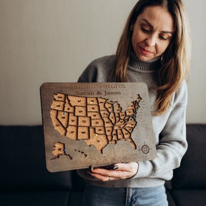 May include: A woman is holding a wooden map of the United States with corkboard states. The map is titled "Our adventures Sarah & Jason" and has push pins in various states.