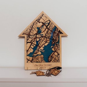 May include: A house-shaped wooden wall plaque featuring a laser-cut street map with dark blue water sections. The base includes the engraved text "Our first HOME 2023" and three small metal hooks. Two wooden keychains with map designs are attached.