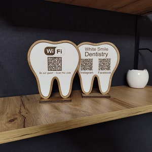 Puede incluir: Dos letreros de madera con forma de diente con fondo blanco. El letrero de la izquierda tiene un símbolo de Wi-Fi y un código QR con el texto "Be our guest - Scan the code". El letrero de la derecha tiene el texto "White Smile Dentistry" y dos códigos QR con el texto "Instagram" y "Facebook".