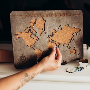 Könnte beinhalten: Eine rechteckige Weltkarte aus Holz mit einer Korkeinlage und dem eingravierten Text Adventure awaits am oberen Rand. Die Karte enthält ein Kompassrosen-Design in der unteren linken Ecke und mehrere kleine bunte Pinnnadeln im Kork.
