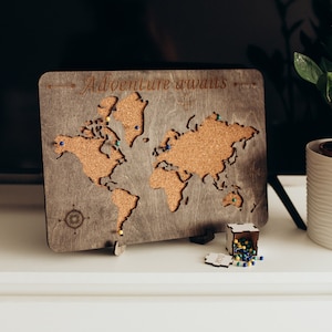 May include: A wooden world map with a cork surface and the text "Adventure awaits" engraved on top. The map is decorated with colourful push pins and a small box of extra pins.