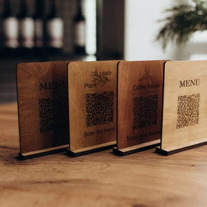 Pode incluir: Quatro placas de mesa retangulares de madeira com bases pretas, cada uma com um código QR gravado a laser. As placas exibem vários textos, incluindo MENU, Plant café e Coffee house, com a instrução Scan for menu ou Scan our menu.