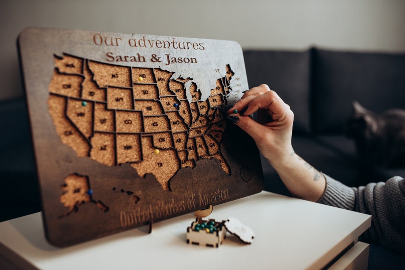 Op de afbeelding: Een rechthoekige houten kaart van de Verenigde Staten met met kurk gevulde staatsuitsparingen. Het bord bevat de tekst Our adventures Sarah & Jason en United States of America. Er zijn diverse gekleurde punaises ingestoken.