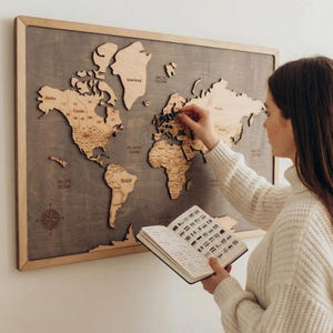 Op de afbeelding: Een rechthoekige houten wereldkaart als wanddecoratie met lichtgekleurde, lasergesneden continenten op een donkergrijze houten achtergrond. De kaart bevat gegraveerde landnamen, een windroos en oceaanlabels. Een klein notitieboek wordt vastgehouden.