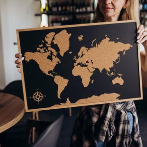 May include: A rectangular world map made of cork on a black background, framed in wood. The continents are in a natural cork color, and a compass rose is in the lower left corner. The map is designed for pinning locations.