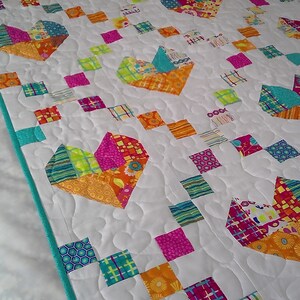 May include: A white quilt with a turquoise border. The quilt is made of patchwork squares in various colours and patterns, including pink, orange, yellow, green, and blue. The squares are arranged in a pattern of hearts.