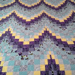 May include: A patchwork quilt featuring squares of yellow, purple, blue and grey fabric. The quilt is made with a variety of patterns and textures.