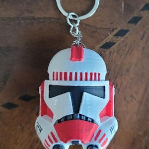 May include: A white and red plastic keychain shaped like a Star Wars clone trooper helmet. The helmet has a black visor and red accents.