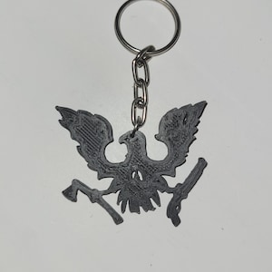 Può includere: Un portachiavi grigio scuro con un emblema di aquila stilizzato con un'ascia e una pistola. Il portachiavi ha un anello e una catena in metallo argentato. Il design dell'aquila è un oggetto stampato in 3D.