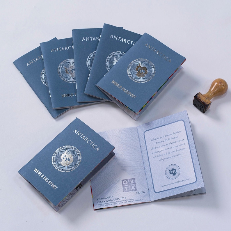 May include: A stack of blue passport-sized booklets with the word "Antarctica" and "World Passport" printed on the cover. The booklets are arranged in a semi-circle on a white surface. A wooden stamp with the word "Antarctica" is visible in the background.