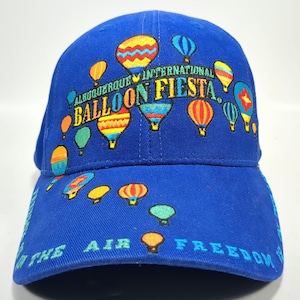 May include: A royal blue baseball cap featuring colorful hot air balloons. The cap is embroidered with the text "Albuquerque International Balloon Fiesta" and "In the Air Freedom". The design includes various sizes and colors of hot air balloons.