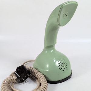 May include: A vintage mint green telephone with a curved handset and a round base. The phone has a coiled cord and a black plug. The base features a speaker grill. The phone is set against a white background.