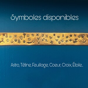 May include: A gold-colored metal strip with various stamped symbols, including zodiac signs, hearts, and stars. The text "Symboles disponibles" is above the strip, and "Astro, T&eacute;tine, Feuillage, Coeur, Croix, &Eacute;toile..." is below it, all against a blue background.