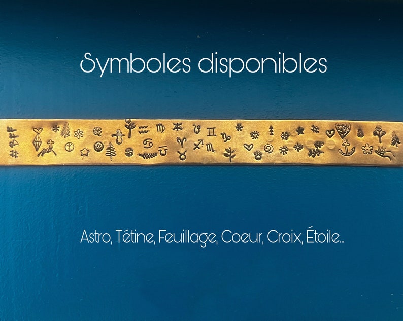 May include: A gold-colored metal strip with various stamped symbols, including zodiac signs, hearts, and stars. The text "Symboles disponibles" is above the strip, and "Astro, T&eacute;tine, Feuillage, Coeur, Croix, &Eacute;toile..." is below it, all against a blue background.