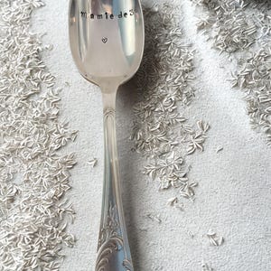 Puede incluir: Una cuchara plateada con las palabras "Mamie de 3" y un pequeño corazón estampado en el cuenco. La cuchara tiene un mango ornamentado con detalles florales. La cuchara está sobre un fondo blanco texturizado.