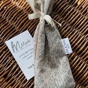 May include: A beige fabric bag with a leaf pattern, tied with a cream-colored ribbon, rests on a woven basket. A small card with the word "Merci" is next to the bag. The bag has a tag that says "Mat & Romy".