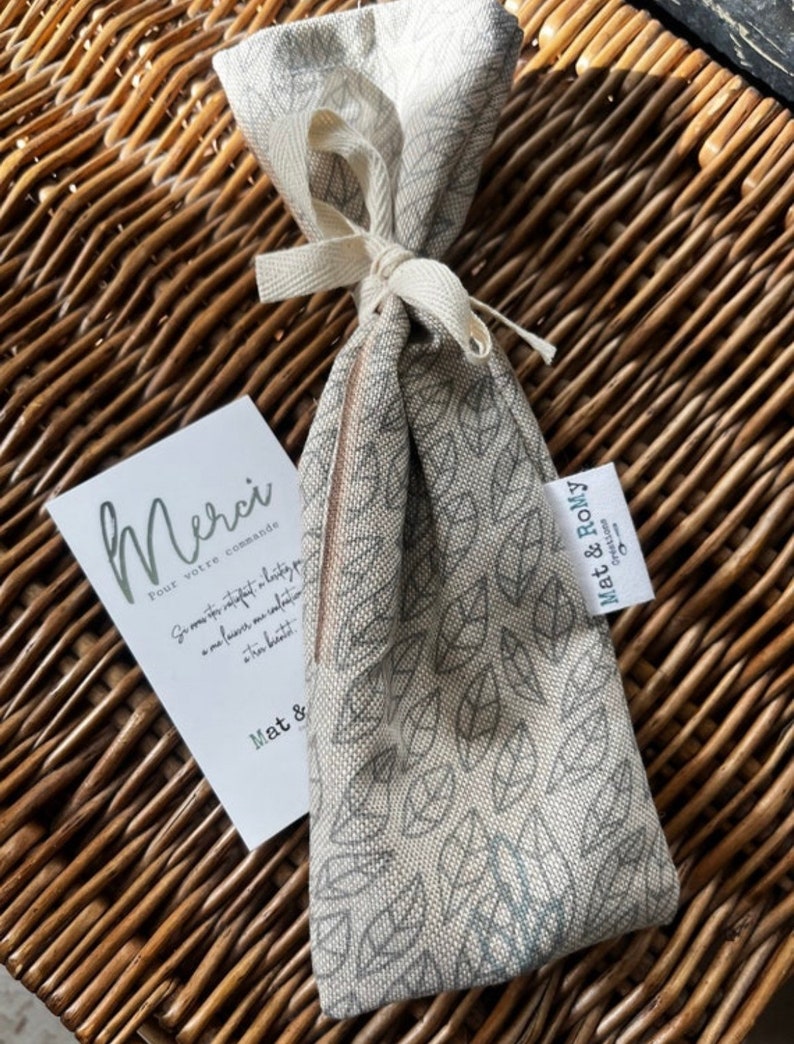 May include: A beige fabric bag with a leaf pattern, tied with a cream-colored ribbon, rests on a woven basket. A small card with the word "Merci" is next to the bag. The bag has a tag that says "Mat & Romy".