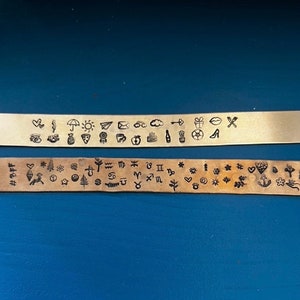 May include: Two metal strips with stamped designs. The top strip is a light gold color and features various small symbols, including a heart, umbrella, pizza slice, and cloud. The bottom strip is a darker bronze color and has zodiac signs and other symbols.