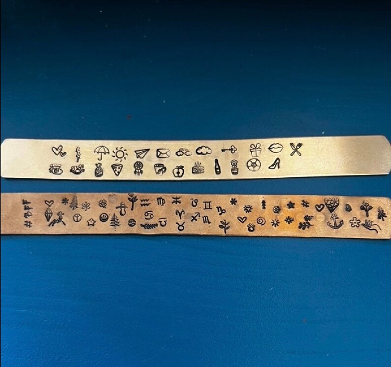 May include: Two metal strips with stamped designs. The top strip is a light gold color and features various small symbols, including a heart, umbrella, pizza slice, and cloud. The bottom strip is a darker bronze color and has zodiac signs and other symbols.
