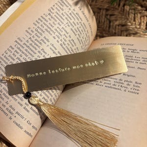 Puede incluir: Un marcapáginas de oro con un corazón y el texto "Bonne lecture mon obet" grabado en él. El marcapáginas tiene un borla.