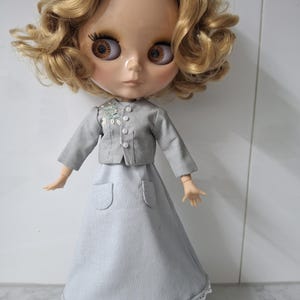 May include: A doll with large brown eyes and curly blonde hair, dressed in a grey jacket and skirt. The jacket features small white buttons and a floral detail. The skirt has pockets and a lace trim. The doll is wearing brown shoes.