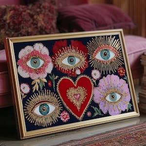 Puede incluir: Una obra de arte enmarcada con flores, corazones y ojos bordados con iris azules. El bordado está detallado con hilo dorado sobre un fondo azul marino, en un marco dorado. La obra se exhibe sobre una superficie rosa.