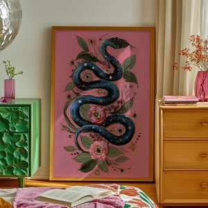 Puede incluir: Una impresión artística enmarcada que presenta una serpiente azul estilizada con detalles de estrellas y oro, rodeada de flores y follaje, sobre un fondo rosa. La obra de arte se exhibe en una habitación con un mueble verde, una cama y una mesita de noche de madera.